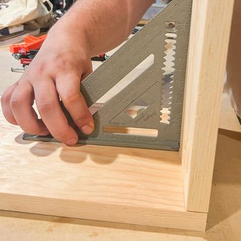 A hand holds a framing square against a piece of wood, positioning it for precise measurement within a workshop environment filled with tools and materials.