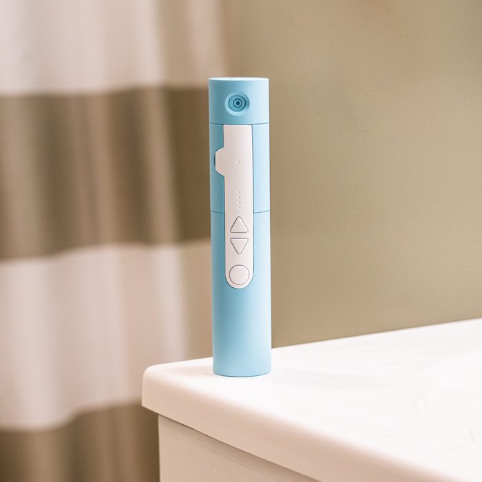 A blue electronic device stands on a bathroom countertop, featuring buttons and indicators, with a blurred striped shower curtain in the background.