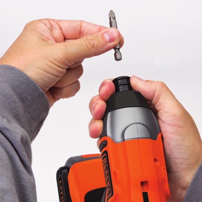 A hand is inserting a screwdriver bit into a power drill, which is held securely in another hand. The background is plain and neutral.