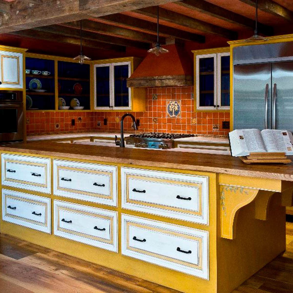 A wooden kitchen island with drawers supports an open cookbook, while cooking appliances and colorful tiled walls create a warm, inviting atmosphere.