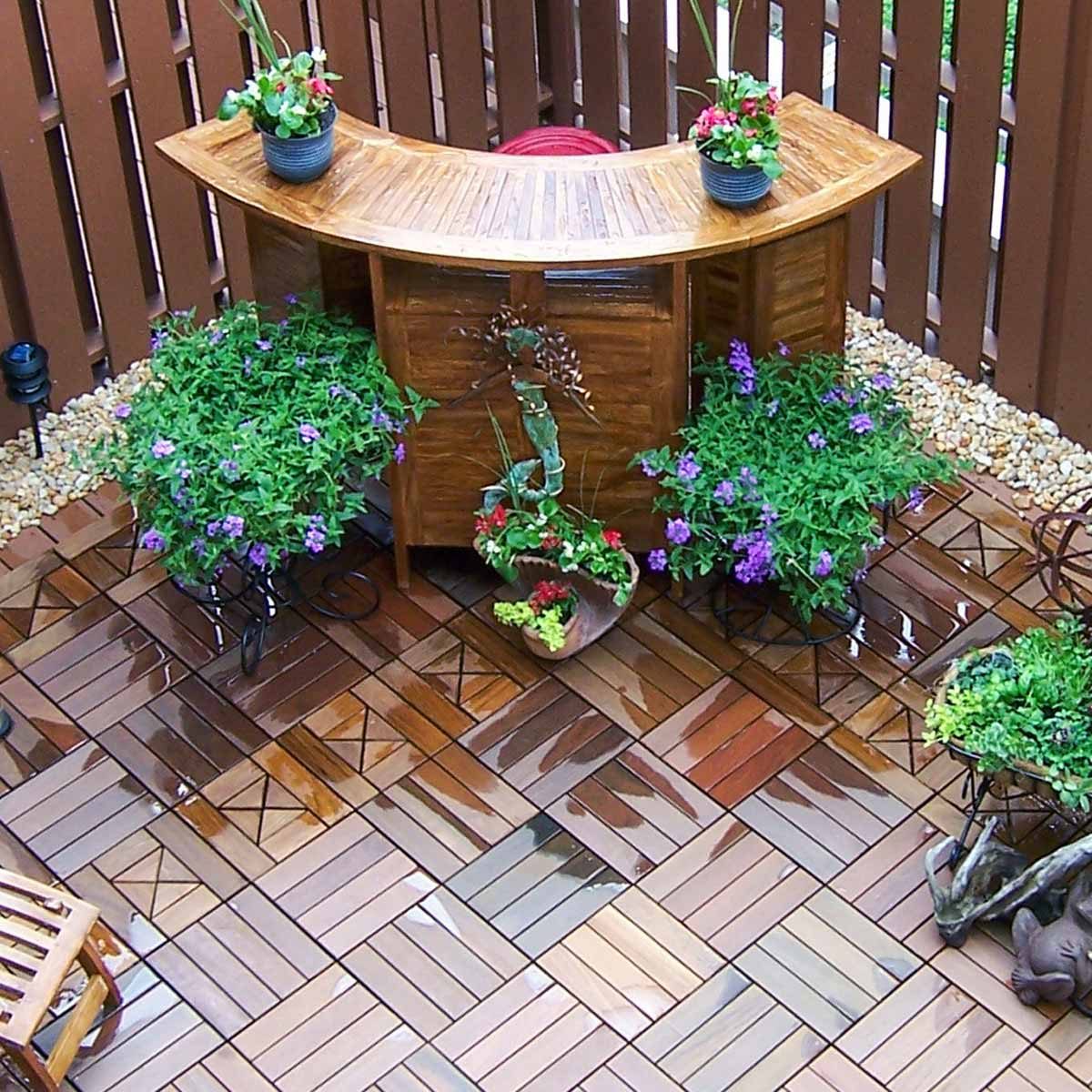 A curved wooden bar sits surrounded by vibrant potted plants on a glossy tiled patio, with decorative stones and a wooden chair nearby.