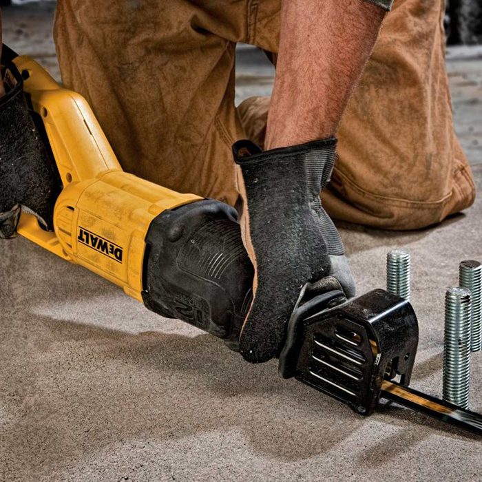 A yellow power tool is being held by a gloved hand, positioned near screws, on a concrete surface in a workshop environment.