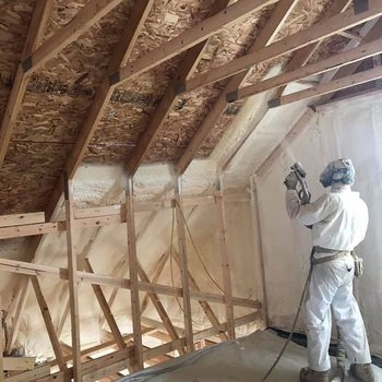 A worker in protective gear sprays insulation on wooden beams in an unfinished attic, surrounded by exposed framing and a partially insulated wall.