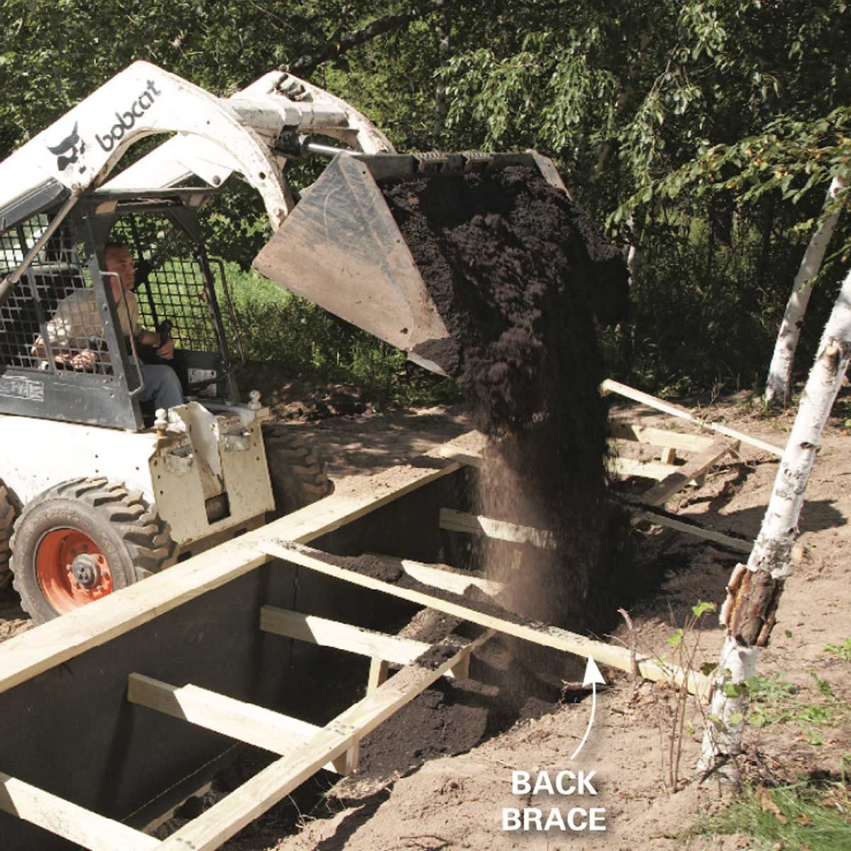 A bobcat loader dumps soil into a dug trench, surrounded by wooden braces and foliage in a sunny outdoor setting.