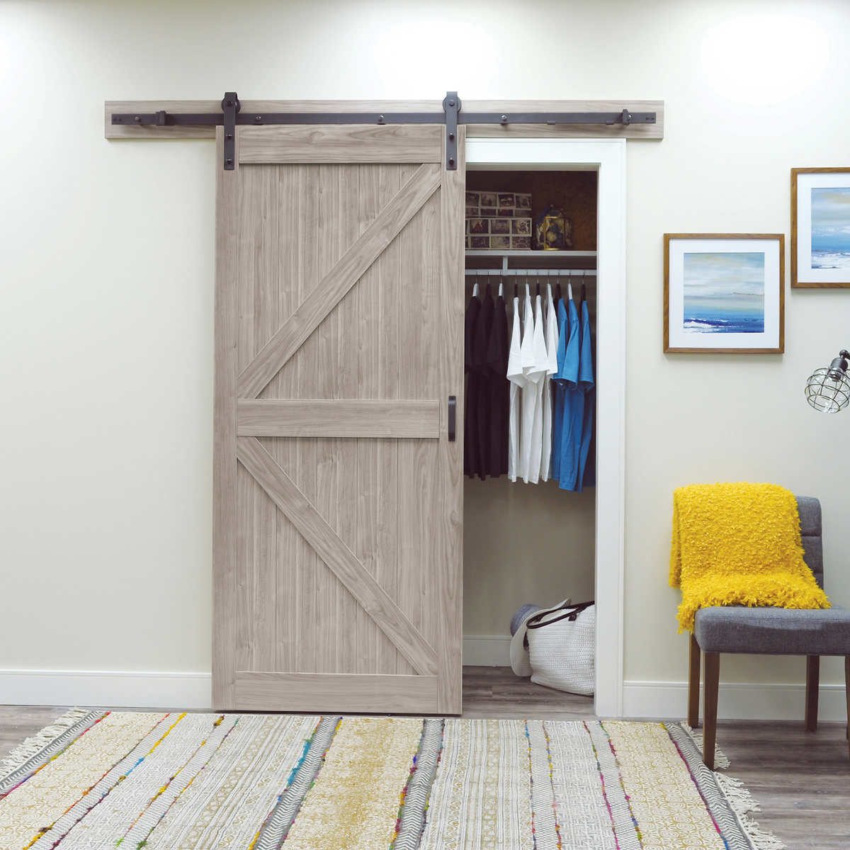 A sliding wooden barn door is partially open, revealing a closet with hanging clothes. A cozy chair and a patterned rug complete the room's decor.