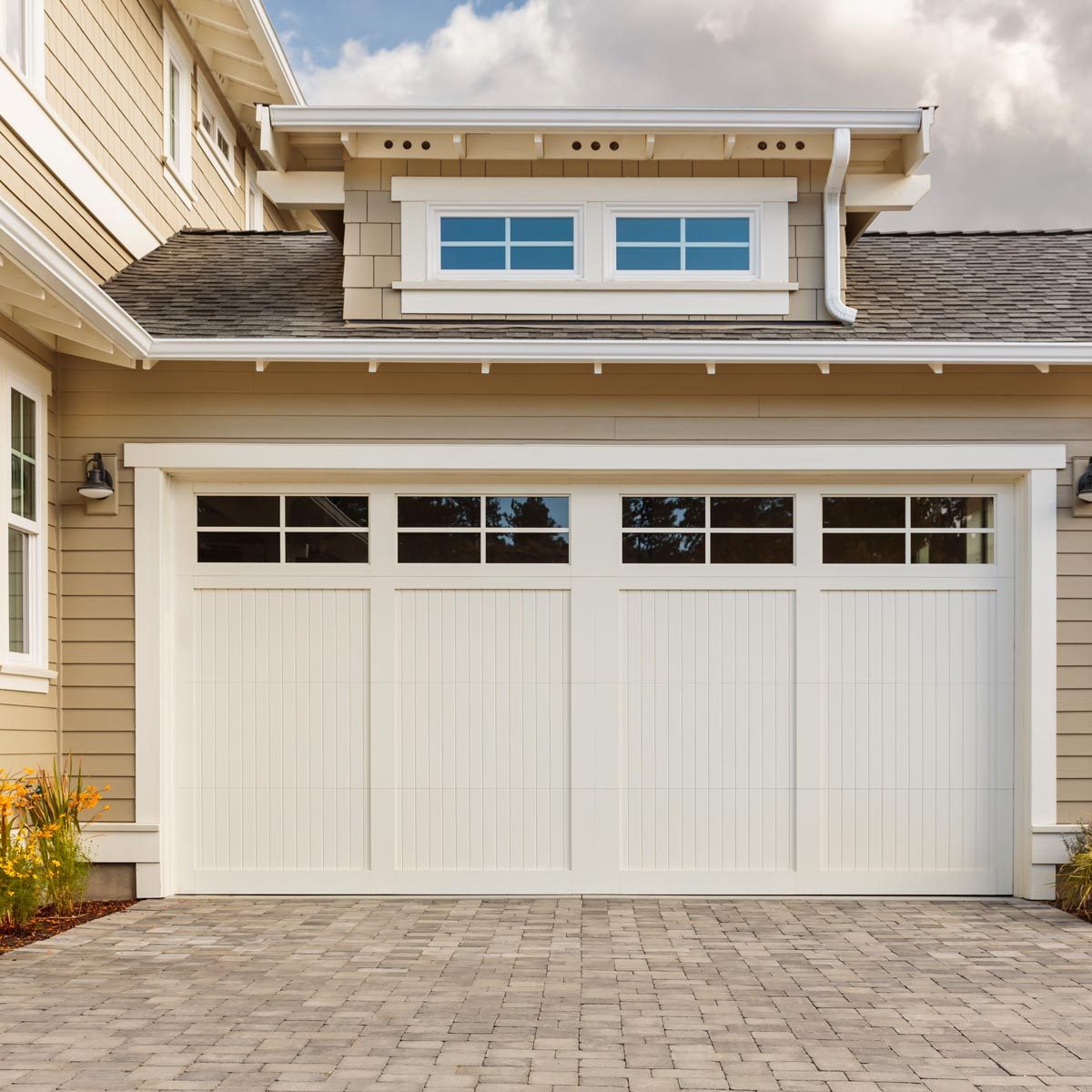 A white garage door occupies the center, slightly open. It's set against a beige house with a small flower bed and paved driveway under a partly cloudy sky.