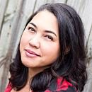 A woman with dark, wavy hair smiles softly while leaning against a wooden fence, wearing a red top. Natural light highlights her features.