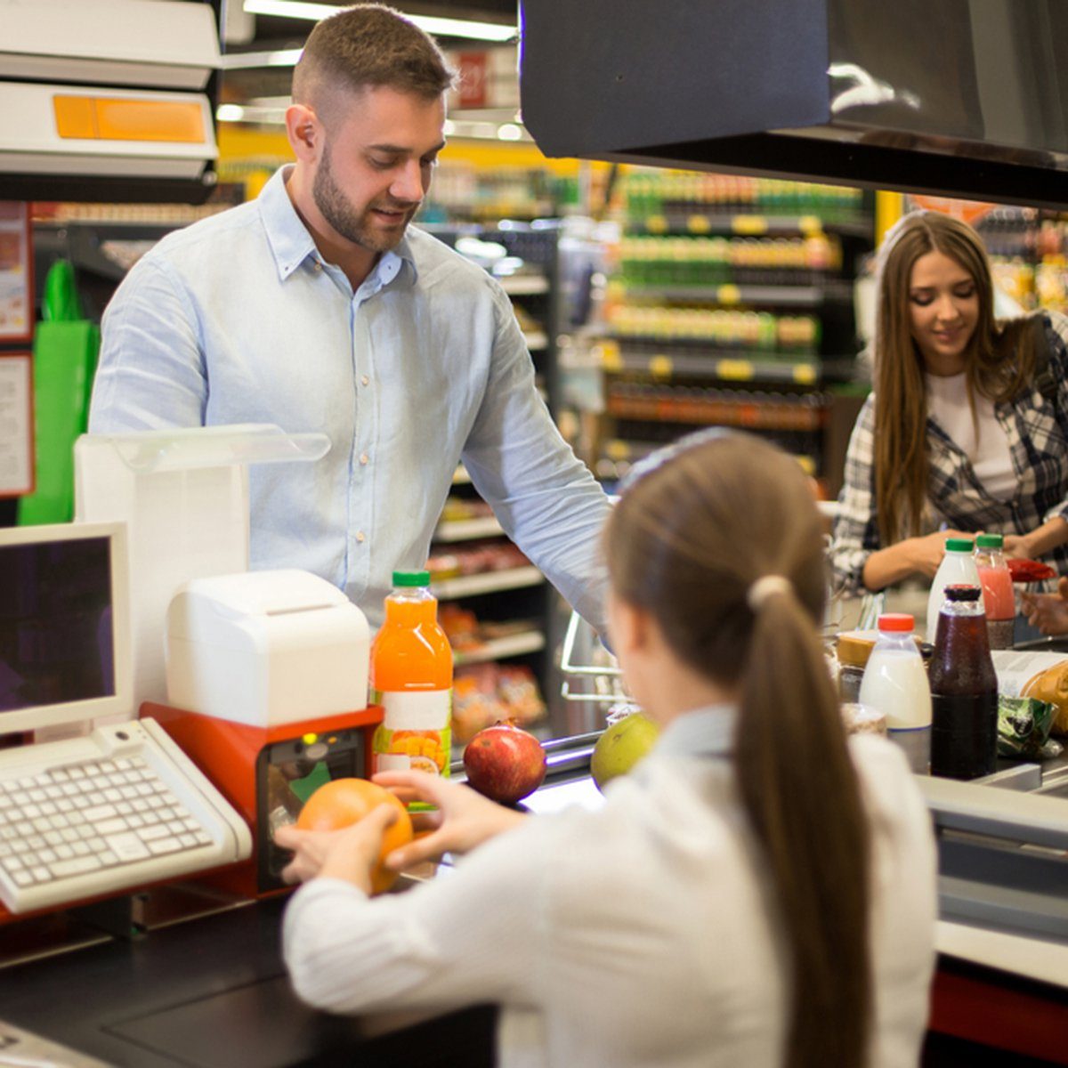 A cashier assists a customer bagging groceries at a supermarket checkout, surrounded by shelves stocked with various products.