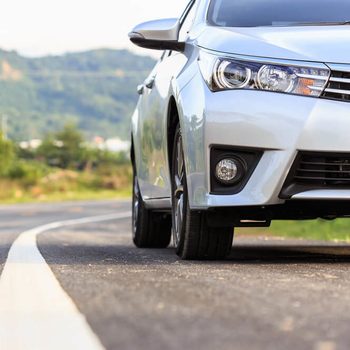 A silver car is parked on a curved road, set against a backdrop of hills and greenery under a clear sky.