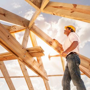 A construction worker uses a hammer to secure wooden beams while standing on a partially constructed roof under a bright sky with scattered clouds.