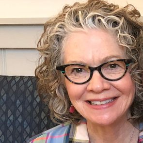 A smiling woman with curly gray hair wears glasses and colorful earrings, seated against a patterned dark background, exuding warmth and friendliness.