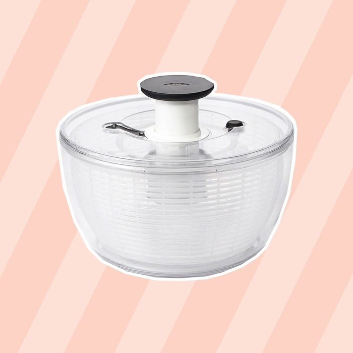 A clear salad spinner uses a central mechanism for drying greens, placed against a soft, striped pastel background.