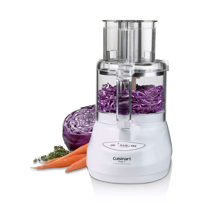 A food processor processes shredded purple cabbage, surrounded by a halved cabbage, carrots, and herbs on a white countertop, demonstrating kitchen preparation.