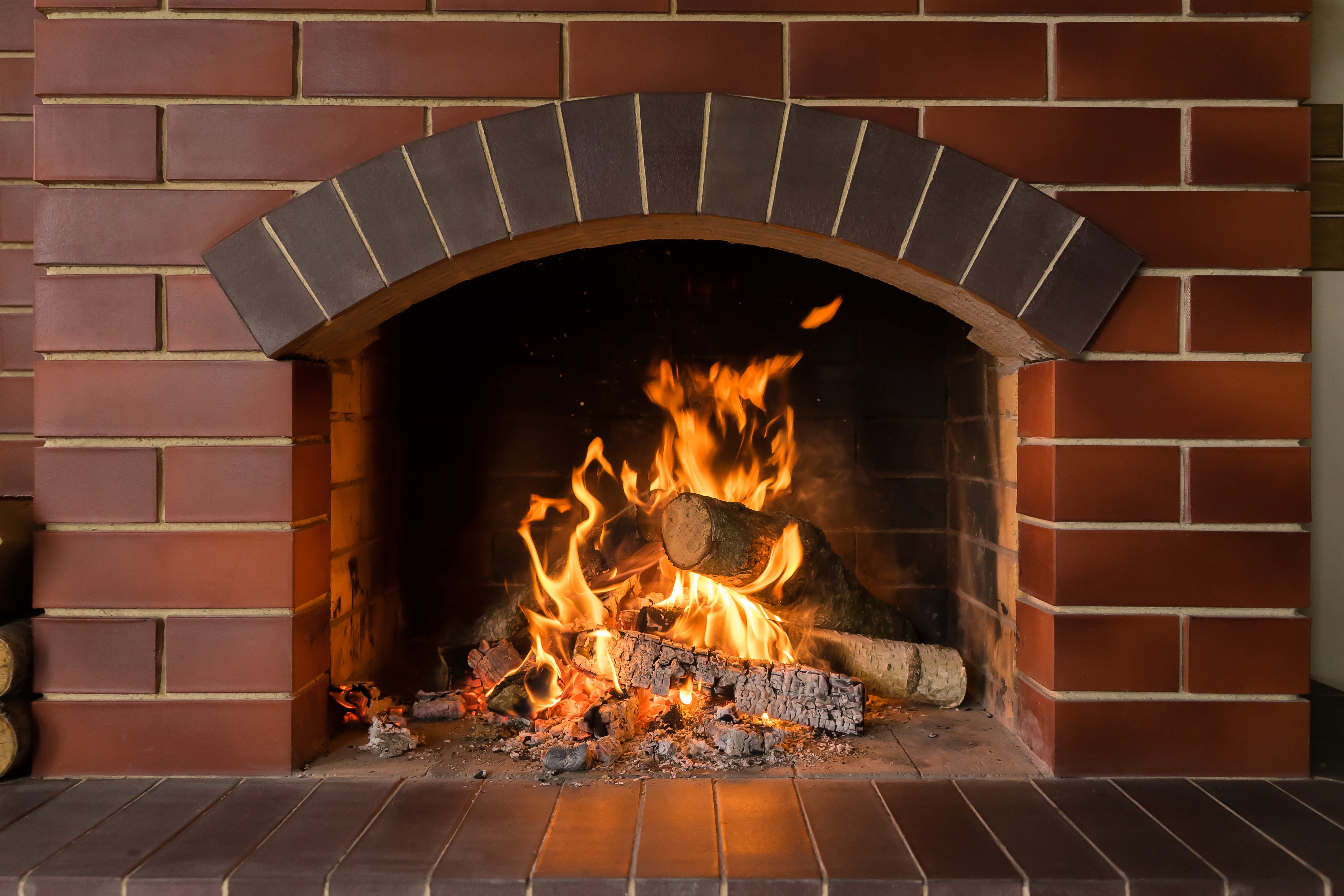 A crackling fire burns brightly in a brick fireplace, surrounded by logs and ashes, creating a warm, inviting atmosphere in a cozy interior.