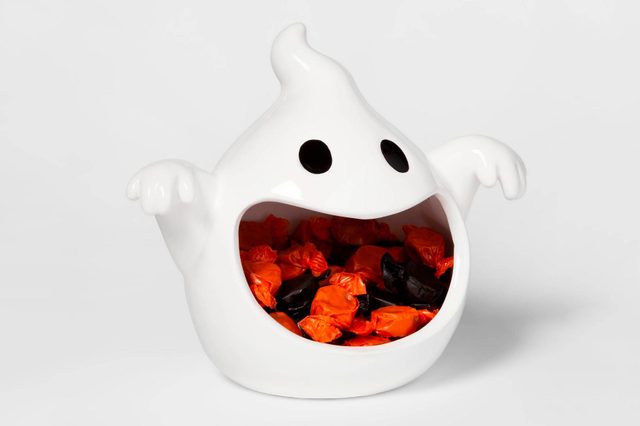 A white ghost-shaped container holds various orange and black wrapped candies, appearing cheerful and festive against a plain background.