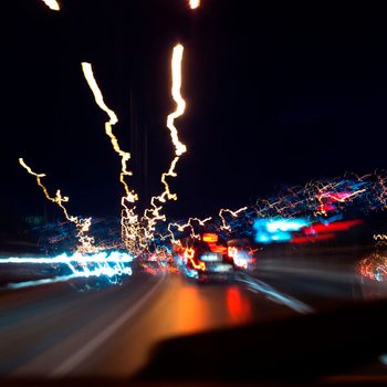 Bright lights streak across a dark roadway, suggesting high speed as vehicles move quickly through a blurred urban environment at night.