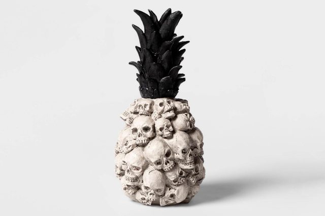A decorative pineapple sculpture, featuring a textured surface of skulls, topped with dark leaves, stands against a plain gray background.