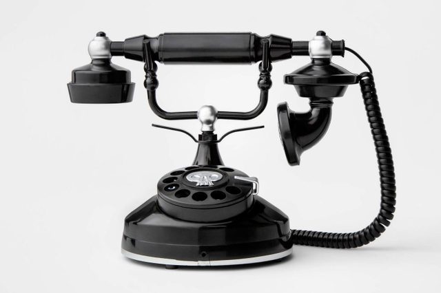 A vintage black rotary phone sits upright, with its handset lifted. It rests on a plain white background, showcasing its retro design.