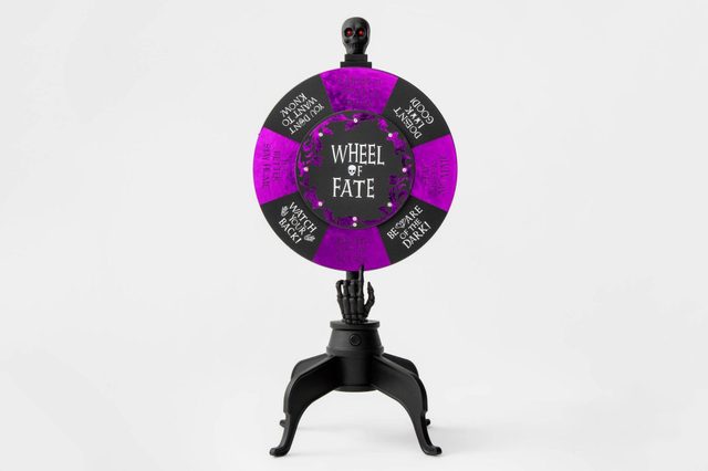A colorful spinning wheel labeled "Wheel of Fate" stands on a black base, with a skeleton hand pointing to various ominous messages.