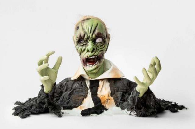 A grotesque green zombie doll with a torn black suit raises its arms, displaying a fierce expression, against a plain white background.