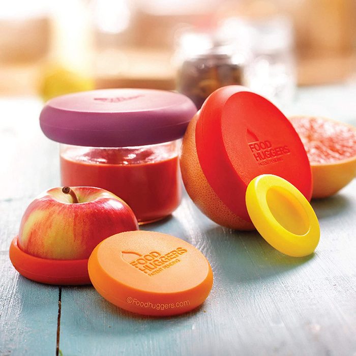 Colorful silicone food huggers surround an apple and a jar of red sauce on a light wooden surface, illustrating food preservation and storage.