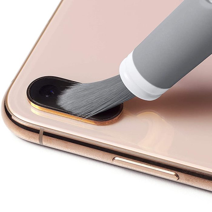 A brush is cleaning a camera lens on a smartphone. The phone is light-colored with a shiny surface and a visible edge.