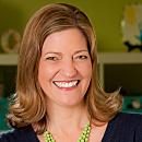 A woman with brown hair smiles brightly while wearing a dark shirt and a green necklace. The background features shelves with colorful objects and a green wall.