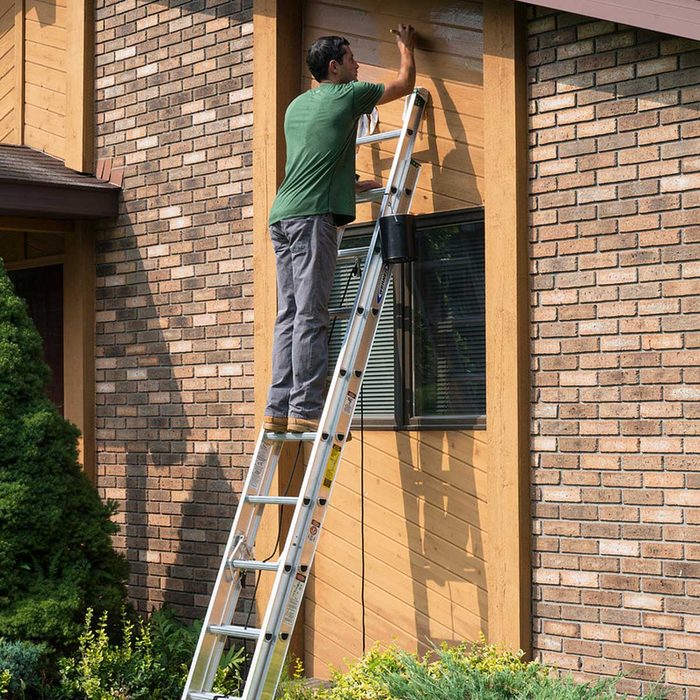 A man stands on a ladder, painting the exterior wall of a house, surrounded by greenery and a brick facade.