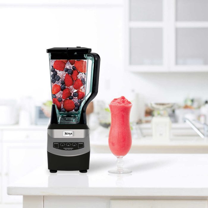 A blender with mixed berries and ice blends, while a pink smoothie sits in a glass on a kitchen counter, surrounded by a bright, clean environment.