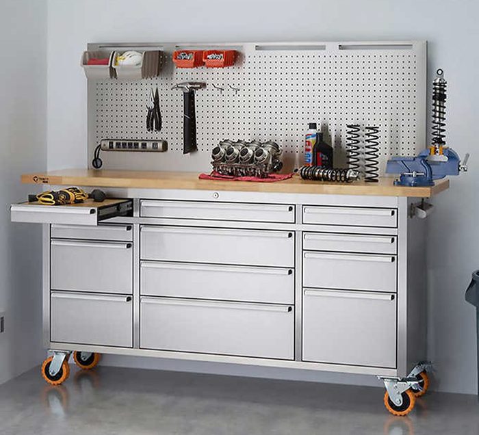 A workshop table with tools, a motor part, and storage drawers, situated against a wall covered in pegboard for organized hanging tools and supplies.