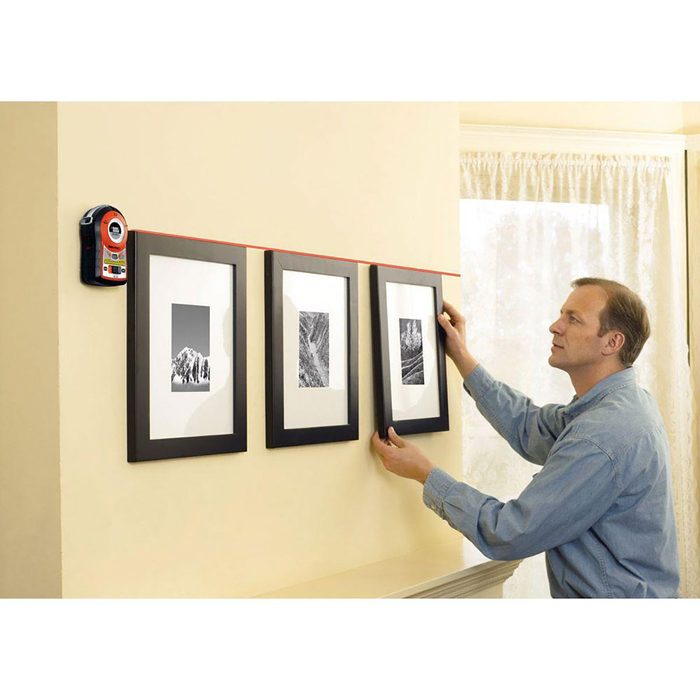 A man adjusts three framed pictures on a wall using a laser level, ensuring they are aligned horizontally in a well-lit room with curtains.