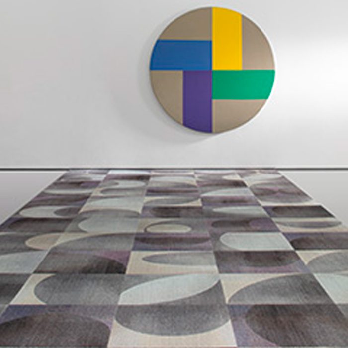 A circular artwork hangs on a wall above a patterned rug. The rug features irregular shapes in various muted colors, creating a contemporary artistic atmosphere.