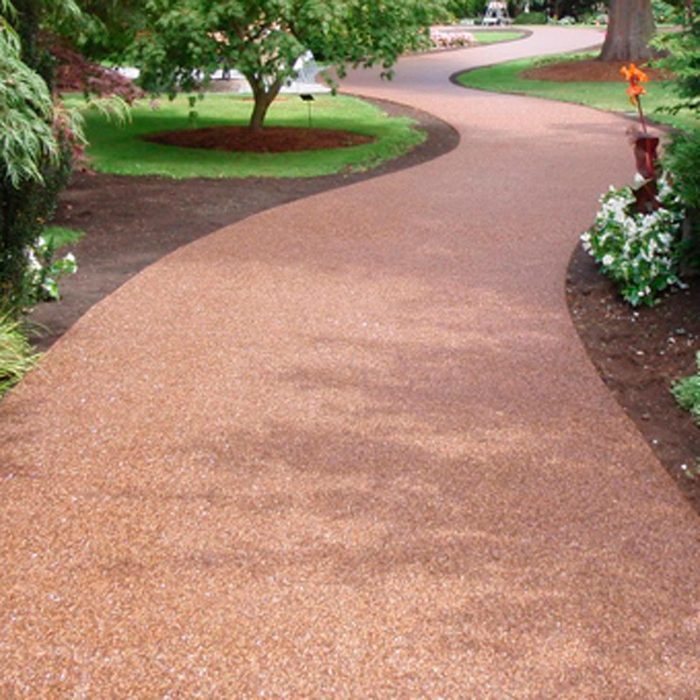 A winding, textured pathway leads through a garden, surrounded by greenery and flowerbeds, inviting leisurely strolls amidst nature.