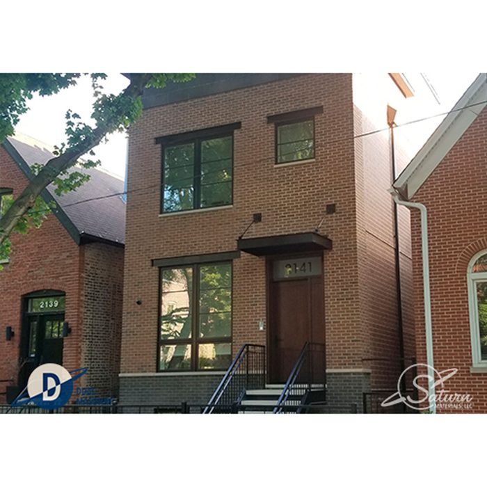 A modern, two-story brick house features large windows and a wooden front door. Surrounding trees provide shade, and nearby houses are visible in the background.