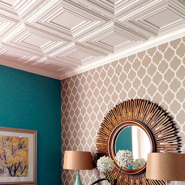 A ceiling features decorative molding. A mirror with a sunburst frame reflects surroundings, alongside two lamps and a vase with flowers against patterned wallpaper.