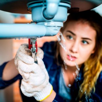 A person wearing gloves adjusts plumbing under a sink, while water leaks from a pipe in a domestic setting.