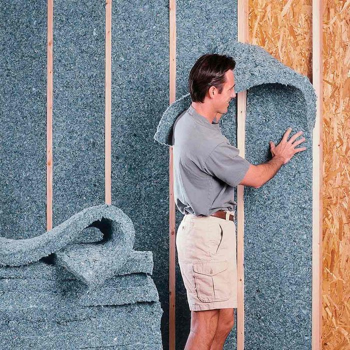 A man installs green insulation material against wooden wall studs, while additional insulation rolls are stacked nearby in a construction setting.