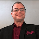 A man in a suit is smiling confidently against a plain background, presenting a professional appearance. He wears glasses and a patterned pocket square.