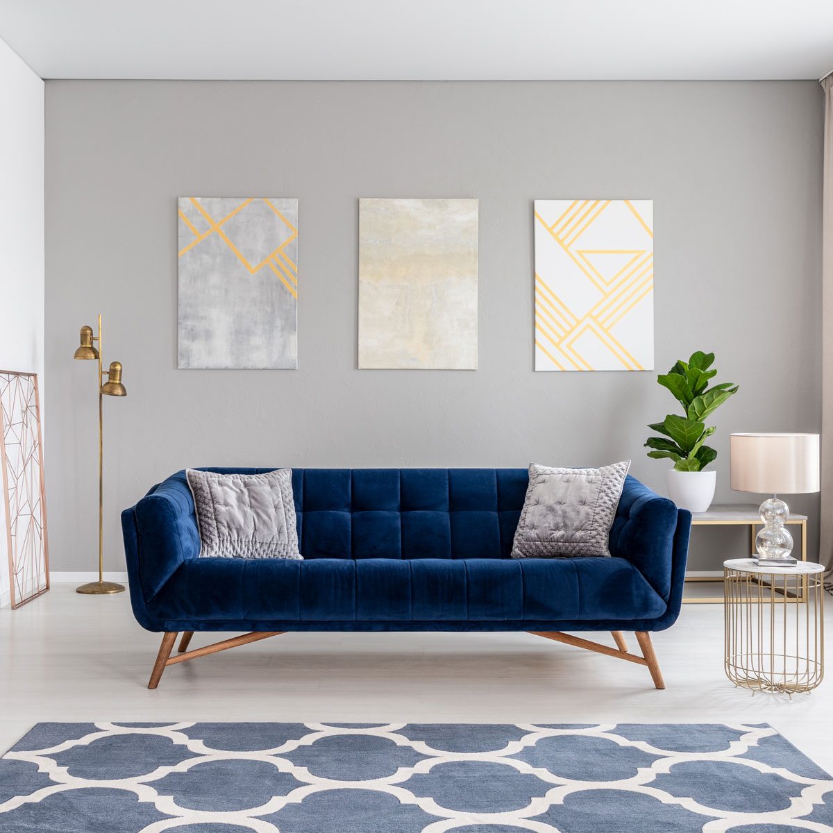 navy blue sofa in the middle of a bright living room interior with gold metal side tables and three paintings on a gray wall couch pillows
