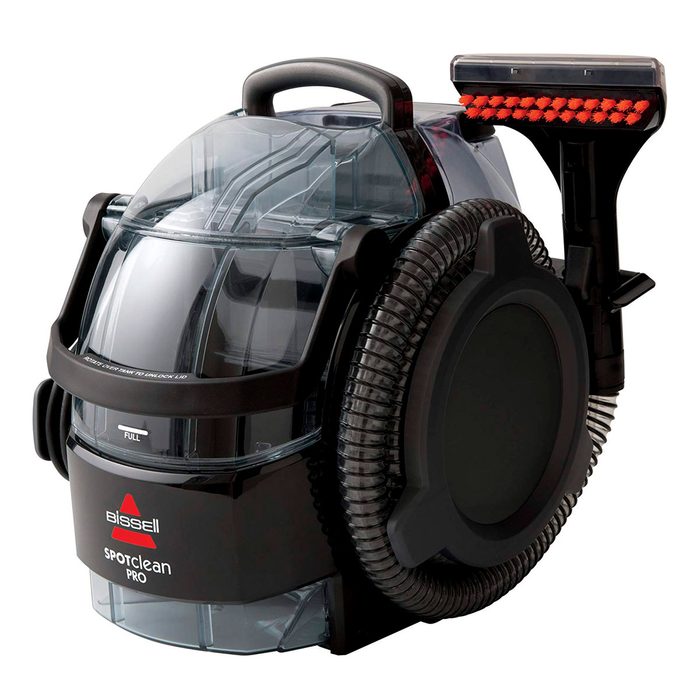 A portable carpet cleaner with a transparent tank and black body is positioned upright, showing its attached cleaning tool and flexible hose for easy maneuvering.