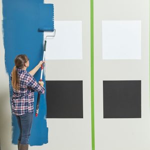 A woman rolls blue paint onto a wall, beside white and black paint samples, with green tape marking off sections in a well-lit room.