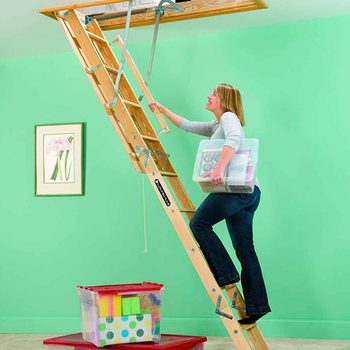 A woman climbs a wooden ladder while holding a storage container, preparing to access a space above in a room with green walls.