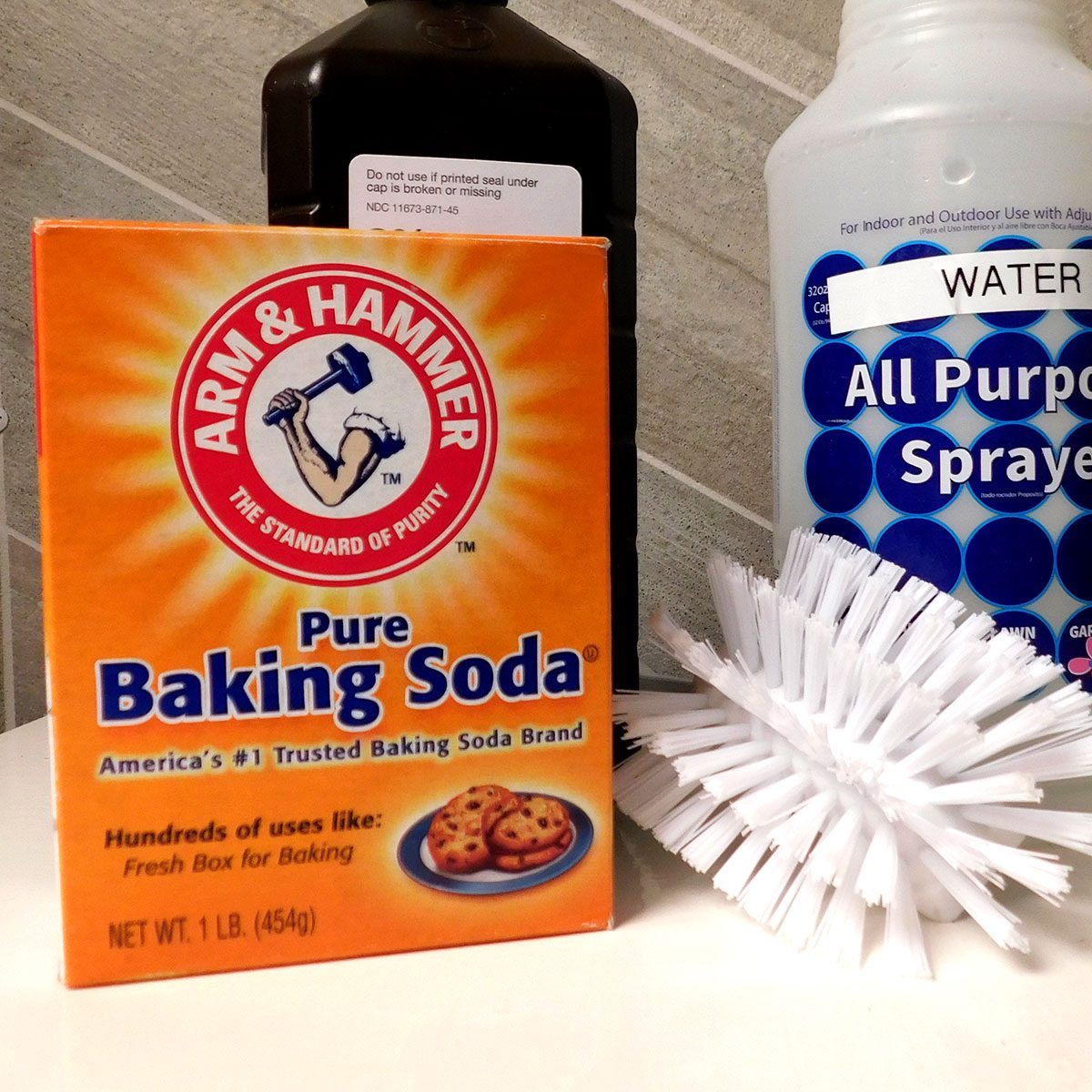 A box of baking soda is positioned beside a black bottle and a white spray bottle, with a cleaning brush leaning against them on a countertop.