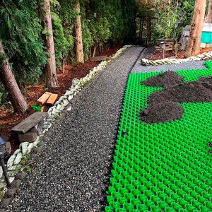 A green, modular paving surface lays beside a gravel pathway, with mounds of soil partially covering the green sections, surrounded by trees and natural debris.