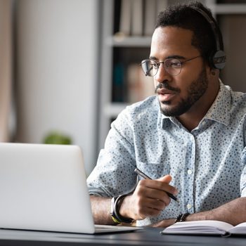 A man wearing headphones works intently at a laptop, taking notes with a pen in a well-lit, stylish workspace.