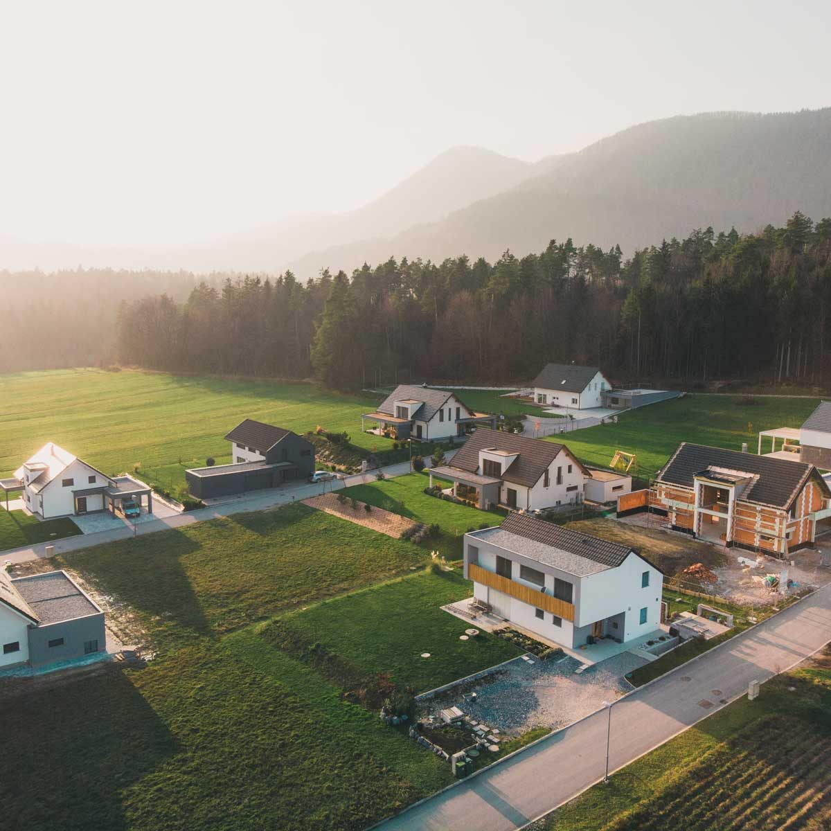 Aerial view of several modern houses arranged in a green field, surrounded by trees and distant mountains, under soft, golden sunlight.