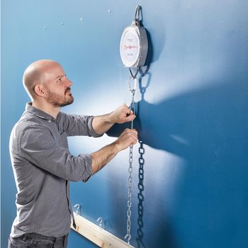 A man adjusts a chain connected to a weighing scale mounted on a blue wall while focused on the measurement process.