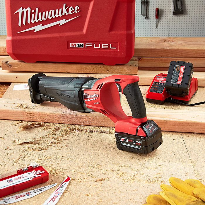A cordless reciprocating saw rests on a work surface, surrounded by wood shavings, a battery charger, and other tools, indicating active woodworking or construction work.