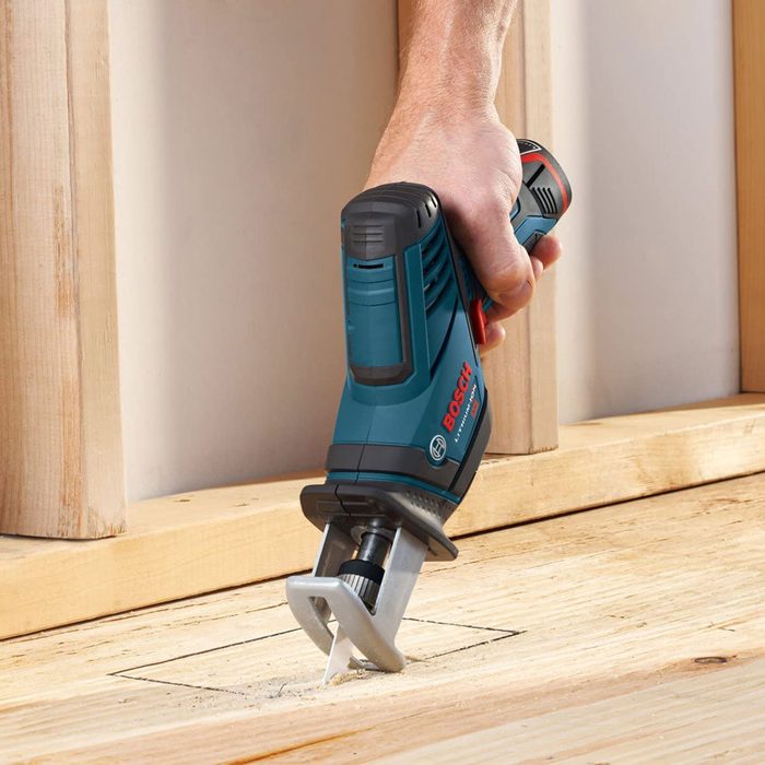 A hand holds a blue and black power tool, cutting into a wooden surface. The environment features unfinished wood surfaces and a light backdrop.
