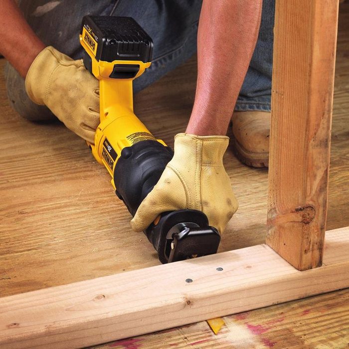 A person wearing gloves uses a power tool to cut through wood on a construction site, surrounded by a wooden floor and framework.
